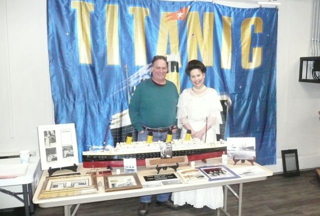 Photo showing Mrs Thomas Andrews and a man standing in front of a large blue Titanic banner, with a model of the Titanic in front of them